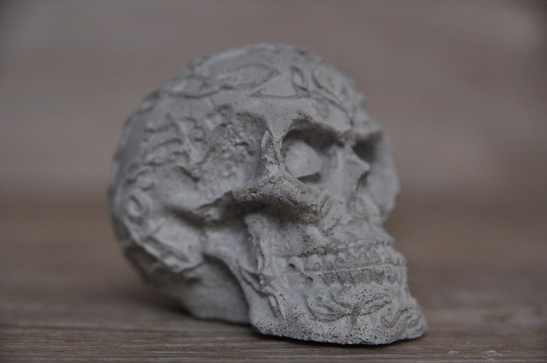May include: A detailed, gray, concrete skull sculpture with intricate carvings and a textured surface.
