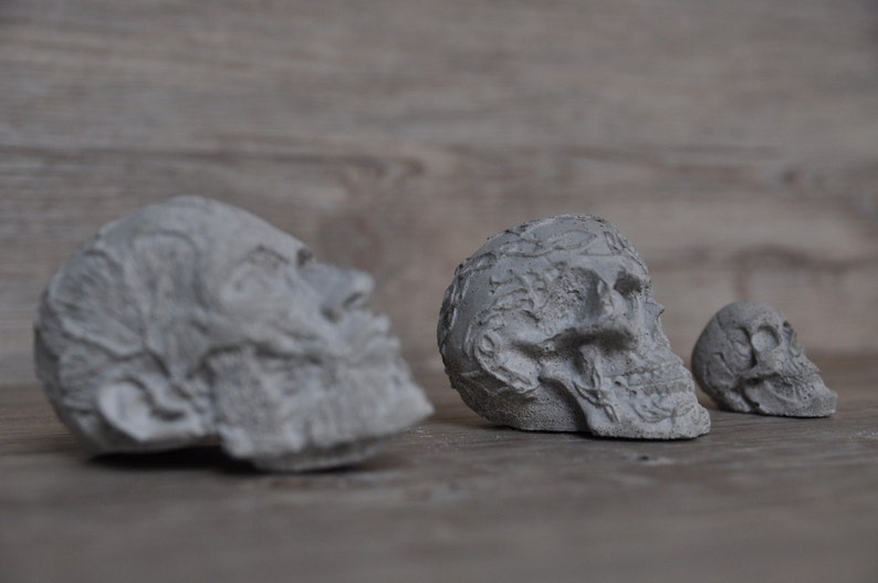 May include: Three gray concrete sculptures of human skulls. The sculptures are arranged in a row from left to right, with the largest sculpture on the left and the smallest sculpture on the right.