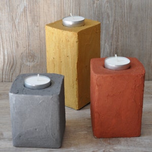 May include: Three concrete candle holders in different colors: gray, yellow, and orange. Each holder has a small, round indentation for a tea light candle.