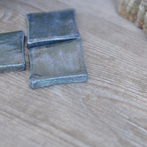 Könnte beinhalten: Vier quadratische Keramikuntersetzer mit blau-grüner Glasur. Die Untersetzer sind auf einer hellen Holzoberfläche angeordnet. Die Kanten der Untersetzer haben eine leicht unregelmäßige Form.