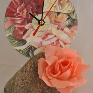 Puede incluir: Un reloj con un diseño floral en una cara redonda, con manecillas negras y una manecilla de segundos roja. El reloj está sentado sobre una roca marrón con una rosa rosa en la parte superior.
