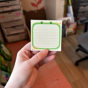 May include: A green frog-shaped sticky note with lined paper and musical notes on the top right corner. The sticky note is being held by a hand.