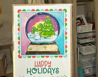May include: A holiday greeting card featuring a snow globe design with a snowman and Christmas trees. The card has a colorful border and the words "HAPPY HOLIDAYS" in green and red. The card is held in a person's hand.