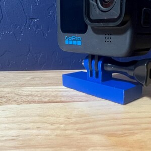 Könnte beinhalten: Eine schwarze GoPro Hero 11 Black Actionkamera, die auf einem blauen Stativ befestigt ist. Die Kamera ist nach vorne gerichtet und das Objektiv ist sichtbar.
