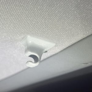 Può includere: Un gancio di plastica bianco, probabilmente per l'interno di un'auto, è montato su una superficie chiara e strutturata. Il gancio ha un design curvo e aperto, ed è posizionato vicino a un bordo grigio liscio.