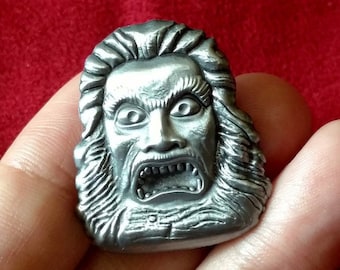 May include: A silver-colored metal pin featuring a detailed, sculpted face with a long, flowing beard and an open mouth, revealing sharp teeth. The face has a menacing expression.