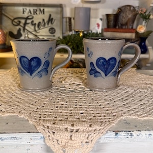 May include: Two matching stoneware mugs with a speckled light gray finish, each featuring a hand-painted cobalt blue heart design on the front. The mugs have a sturdy handle, a slightly flared rim, and a raised base, showcasing a classic pottery style.