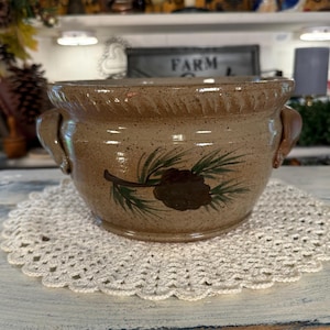 May include: A tan speckled ceramic bowl with two side handles and a decorative rim. The front of the bowl features a hand-painted brown pinecone with green pine needles. The bowl sits on a round, white, crocheted lace doily with a scalloped edge.