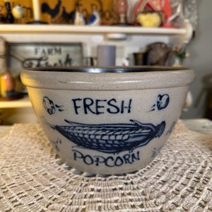 May include: A beige ceramic bowl with a blue rim and blue lettering that reads "FRESH POPCORN". The bowl features a blue corn graphic. The bowl is sitting on a cream-colored, lace doily.