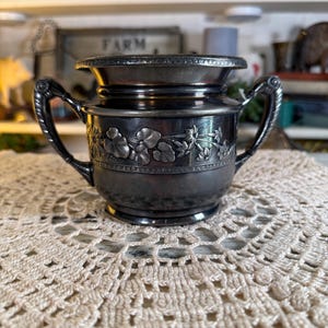 May include: An antique silver-plated sugar bowl with two handles and a lid. The bowl features floral relief detailing around the body. The sugar bowl is sitting on a cream-colored, crocheted doily.