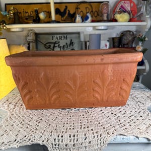 May include: A rectangular terracotta planter with embossed leaf designs. The planter is a warm, earthy orange-brown color and sits on a cream-colored, crocheted doily. The planter is a decorative item for indoor or outdoor use.