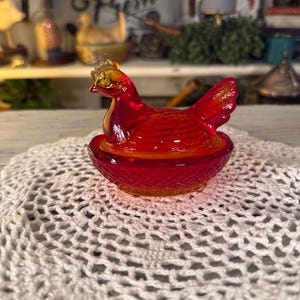 May include: A vibrant red glass hen-shaped trinket box. The hen sits atop a matching nest, with a textured surface. The lid of the hen is detailed with a golden comb. A charming decorative piece for a farmhouse-style home.