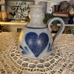 May include: A stoneware pitcher with a blue heart design. The pitcher has a handle and a spout. The background is a shelf with various decorative items. The pitcher is sitting on a cream-colored, crocheted doily.