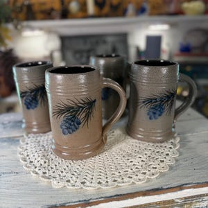 May include: Four stoneware mugs with handles, each featuring a brown and blue pinecone design. The mugs have a textured, speckled finish and a dark interior. They are arranged on a white crocheted doily, creating a rustic, handcrafted aesthetic.