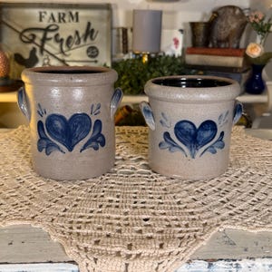 May include: Two cylindrical stoneware crocks with a light tan finish, each featuring a hand-painted blue heart design and two side handles. The taller crock is marked with the number 12 in blue paint on the front, while both have dark rims.