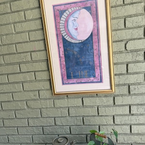 May include: A framed rectangular wall art piece featuring a stylized crescent moon with a human face, colored in shades of blue and pink. The artwork includes the text "WELCOME TO THE MOON" and "LUNE" set against a dark blue background with stars.
