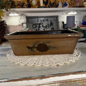 May include: A rectangular ceramic loaf pan with a glossy tan finish and a dark brown interior. The exterior features a hand-painted design of a brown pinecone with green pine needles centered on the front side of the vessel.