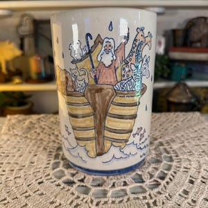 May include: A white ceramic mug with a black handle and the text "But First, Coffee" in a bold, black font. The mug is set against a blurred background, suggesting a cozy setting.