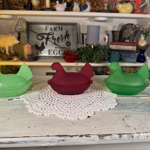 May include: Three glass hen-shaped covered dishes in green and burgundy. The dishes are sitting on a white lace doily. The hen-shaped dishes are a vintage farmhouse style.