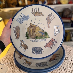 May include: A set of ceramic plates with a blue rim and a cream-colored center. The plates feature illustrations of animals and Noah's Ark, with the text "They went in two and two unto Noah into the ark."