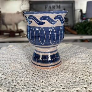 May include: A ceramic goblet featuring a flared rim and a pedestal base. The exterior is decorated with hand-painted blue wave patterns near the top, a central band of white vertical oval outlines on a blue background, and blue scalloped details.