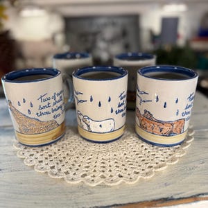 May include: Three ceramic mugs with blue rims and bases, featuring hand-painted illustrations of animals including a giraffe, sheep, and cows. Each mug displays blue text that reads, Two of every sort shall thou bring into the ark.