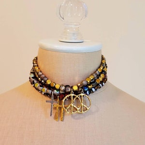 May include: A multi-layered necklace with gold and iridescent beads. The necklace has three charms: a peace sign, a cross, and another peace sign.
