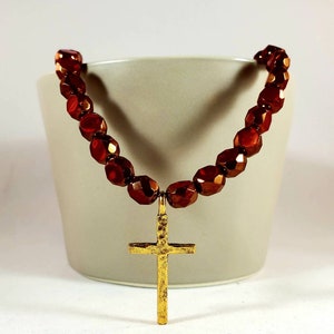 May include: A gold cross pendant necklace with a brown beaded chain. The beads are faceted and have a metallic sheen.