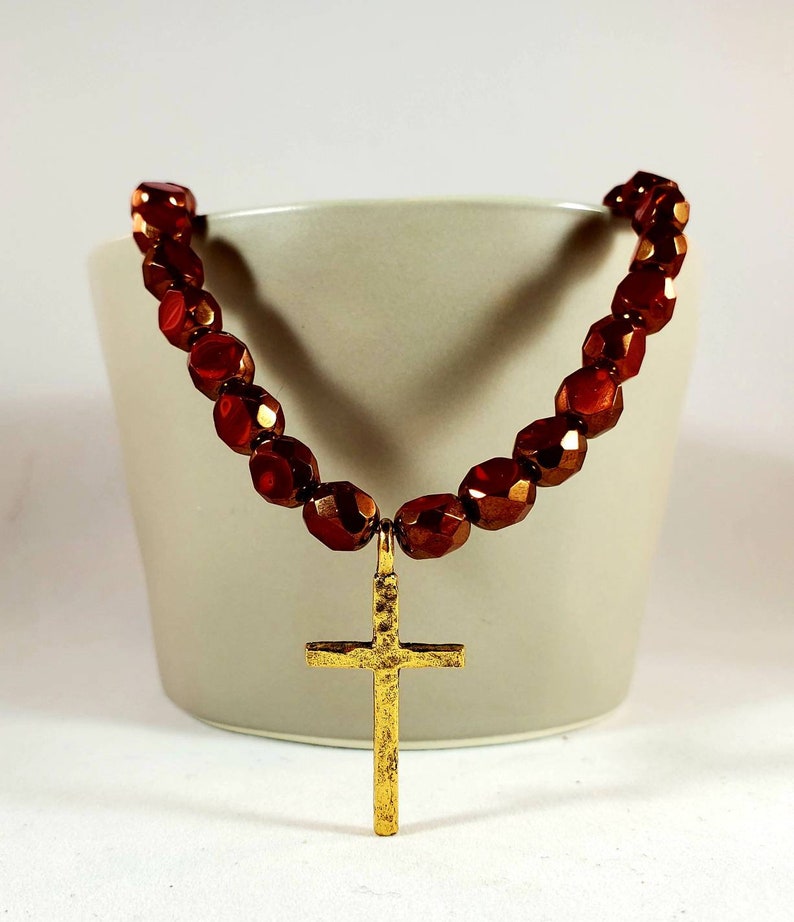 May include: A gold cross pendant necklace with a brown beaded chain. The beads are faceted and have a metallic sheen.