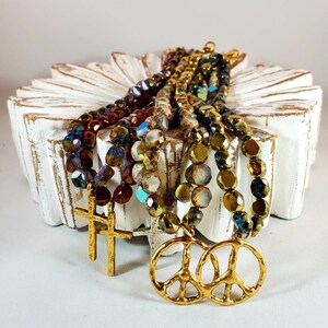 May include: Multi-strand necklace with iridescent glass beads in shades of brown, gold, and blue. The necklace features two gold peace sign charms and two gold cross charms.