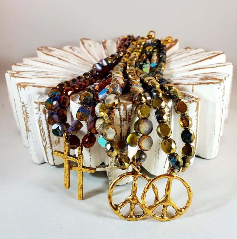 May include: Multi-strand necklace with iridescent glass beads in shades of brown, gold, and blue. The necklace features two gold peace sign charms and two gold cross charms.