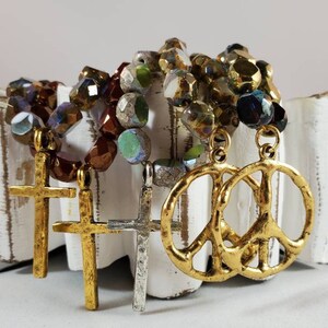 May include: Three gold-tone necklaces with different charms. One necklace has a peace sign charm, and the other two have cross charms. The necklaces are made of iridescent glass beads in shades of brown, green, and blue.