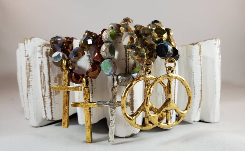 May include: Three gold-tone necklaces with different charms. One necklace has a peace sign charm, and the other two have cross charms. The necklaces are made of iridescent glass beads in shades of brown, green, and blue.