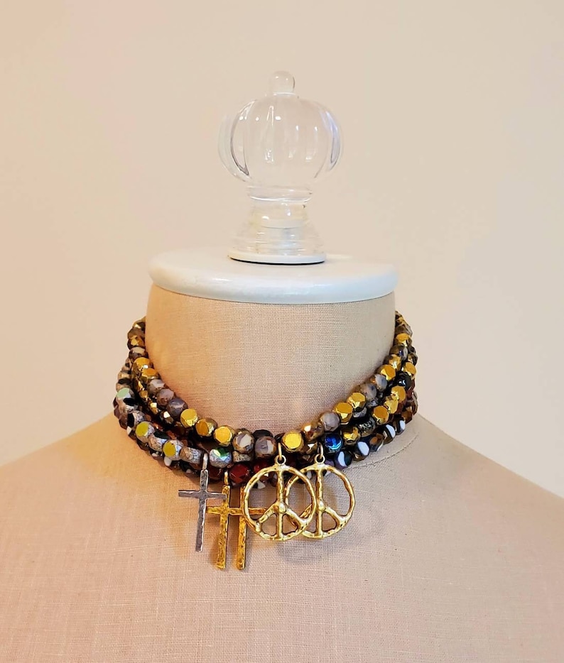 May include: A multi-layered necklace with gold and iridescent beads. The necklace has three charms: a peace sign, a cross, and another peace sign.