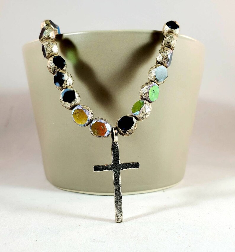 May include: A silver cross pendant necklace with a beaded chain. The beads are iridescent and have a variety of colors, including black, green, yellow, and orange.