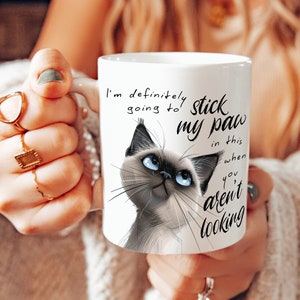May include: White ceramic mug with a Siamese cat illustration and the text "I'm definitely going to stick my paw in this when you aren't looking". The cat has blue eyes. The mug has a curved handle.