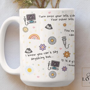 May include: White ceramic mug with a colourful design featuring flowers, rainbows, and ultrasound images. The text on the mug reads "Turn onto your left side Your other left You're can I know you can't say anything but... Is it a gi -ever"
