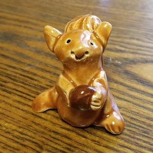 May include: A brown ceramic squirrel figurine holding a brown acorn. The squirrel has a happy expression and is sitting on a wooden surface.