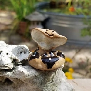 May include: A ceramic snake figurine with a smiling face, tan body, and black and white markings. The snake is perched on a light-colored rock, with a blurred outdoor background.