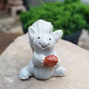May include: A small, gray ceramic squirrel figurine holding a brown acorn. The squirrel has a happy expression and is sitting on a gray rock.
