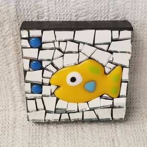 May include: A mosaic art piece featuring a yellow fish with green spots and a blue accent, set against a background of mirrored tiles. Three blue glass bubbles are positioned above the fish. The top edge is black.