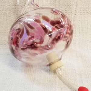 May include: A clear glass essential oil diffuser with a bulbous body and a curved spout. The body is decorated with pink and purple swirls. A cork stopper and a red-tipped glass tube are attached.