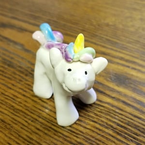 May include: A white ceramic unicorn figurine with a rainbow mane and a yellow horn. The unicorn has a playful expression and is standing on a wooden surface.