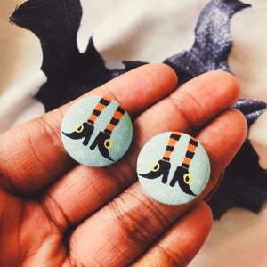 May include: Two round, light blue Halloween-themed buttons featuring witch legs with black and orange striped stockings and black shoes. The buttons are held in a hand, with a bat-shaped decoration in the background.