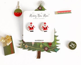 May include: A pair of Santa Claus stud earrings on a white card, with the text "Honey Bea Hive Handmade Accessories". The earrings are red, white, and black. Christmas-themed decorations surround the earrings, including a gift box and a "JOY" ornament.