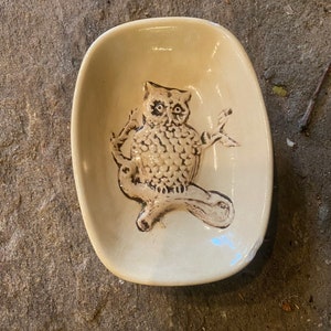 May include: A ceramic dish with a brown owl design. The dish is oval shaped and has a light beige glaze.