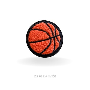 May include: A round, orange basketball patch with black lines and a black border. The patch has a textured, fuzzy appearance. The text "LOLA AND BEAN CREATIONS" is at the bottom of the image.