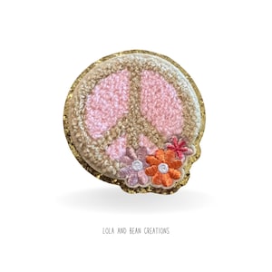 May include: A pink peace sign with a gold glitter outline and embroidered flowers. The flowers are pink, orange, and red.