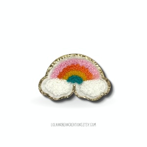 May include: A rainbow-colored felt patch with a gold glitter outline and white felt clouds. The rainbow has pink, orange, yellow, green, and blue stripes.