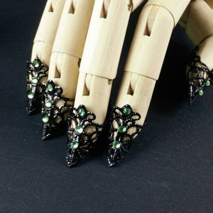 Könnte beinhalten: Schwarze Spitzen-Fingerhut-Ringe mit grünen Strasssteinen. Die Ringe werden an einer Holzhandpuppe getragen.